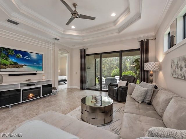 living room with ceiling fan, ornamental molding, a tray ceiling, and a fireplace