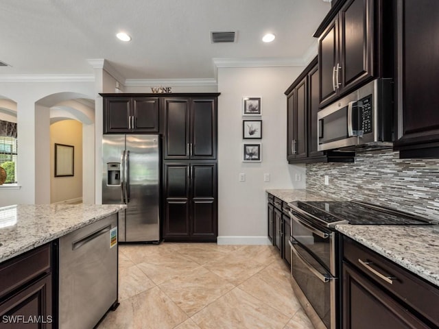 kitchen with dark brown cabinetry, light stone counters, ornamental molding, stainless steel appliances, and backsplash