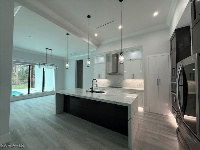 kitchen with stainless steel refrigerator, white cabinetry, wall chimney range hood, a large island with sink, and pendant lighting