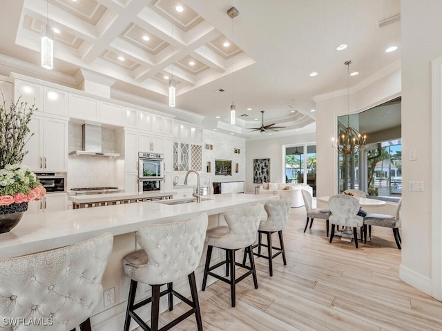 kitchen featuring wall chimney range hood, pendant lighting, a breakfast bar, white cabinets, and ceiling fan with notable chandelier