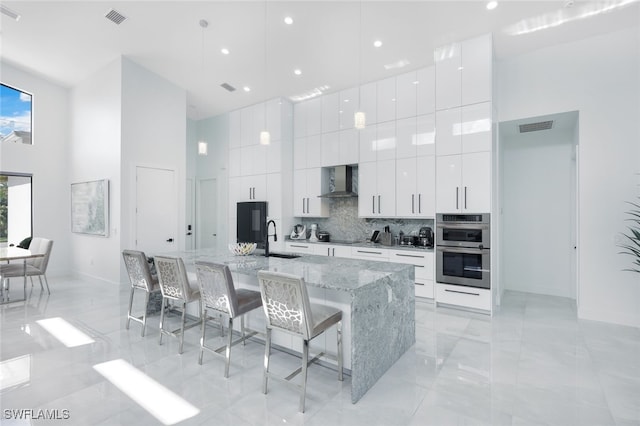 kitchen featuring white cabinets, an island with sink, stainless steel double oven, a breakfast bar area, and a towering ceiling