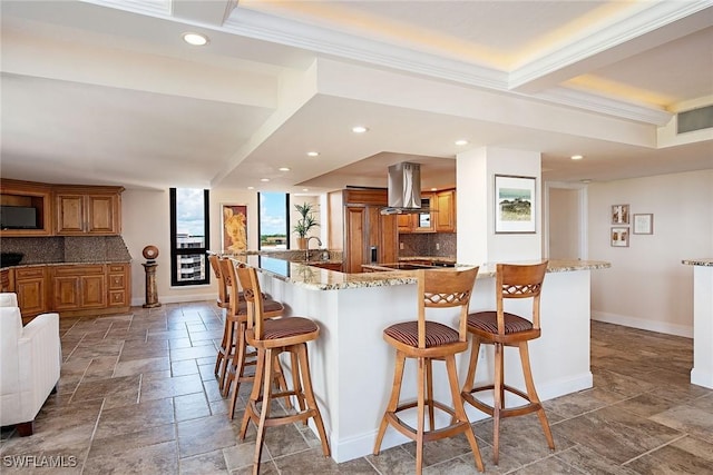 kitchen featuring stone tile floors, baseboards, brown cabinetry, island range hood, and a kitchen breakfast bar