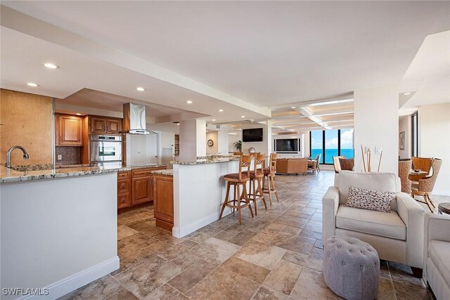 kitchen with beamed ceiling, light stone countertops, island exhaust hood, a kitchen bar, and double oven