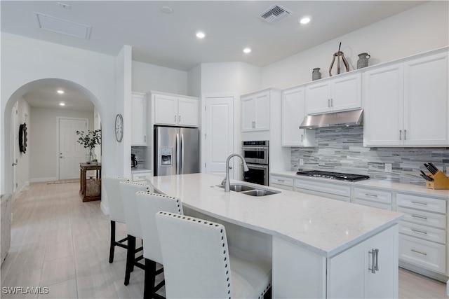 kitchen with arched walkways, under cabinet range hood, a sink, appliances with stainless steel finishes, and decorative backsplash