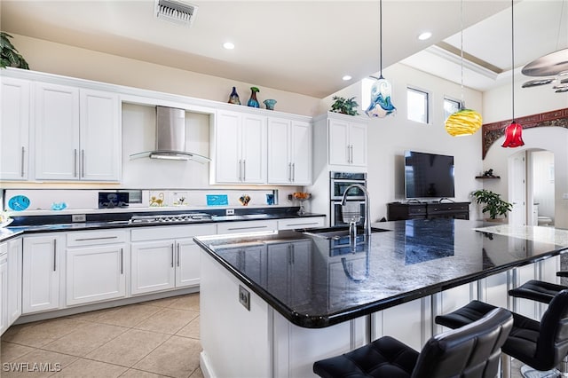 kitchen featuring wall chimney exhaust hood, a kitchen island with sink, a kitchen bar, and decorative light fixtures