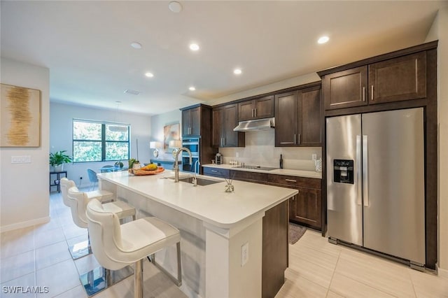 kitchen with dark brown cabinets, under cabinet range hood, an island with sink, stainless steel appliances, and a sink