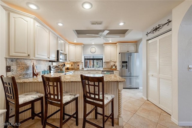 kitchen featuring arched walkways, stainless steel appliances, a peninsula, light stone countertops, and a tray ceiling