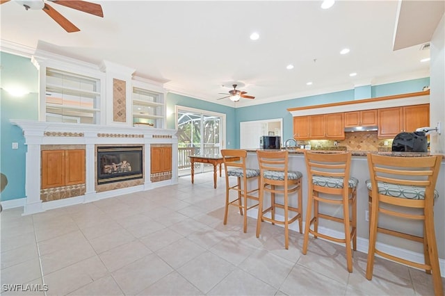 kitchen featuring brown cabinets, crown molding, a ceiling fan, a glass covered fireplace, and under cabinet range hood