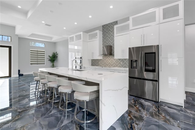 kitchen with white cabinetry, light stone counters, decorative backsplash, stainless steel fridge with ice dispenser, and a kitchen island with sink