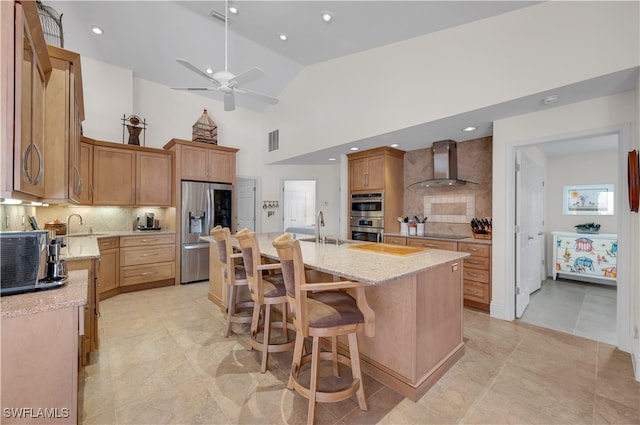 kitchen with light stone countertops, wall chimney exhaust hood, appliances with stainless steel finishes, a kitchen island with sink, and ceiling fan