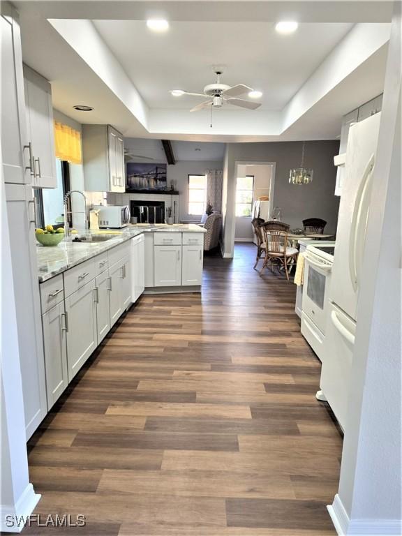 kitchen with white cabinets, ceiling fan with notable chandelier, kitchen peninsula, a raised ceiling, and dark hardwood / wood-style flooring