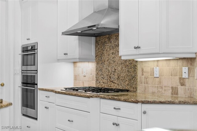 kitchen featuring wall chimney exhaust hood, stainless steel appliances, backsplash, dark stone counters, and white cabinets