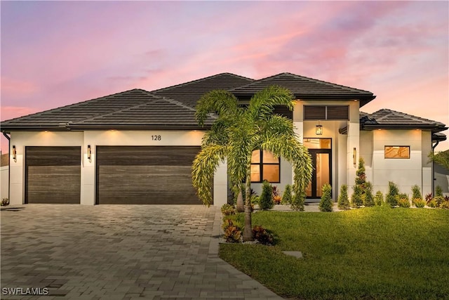 prairie-style home with decorative driveway, a tile roof, stucco siding, an attached garage, and a front yard