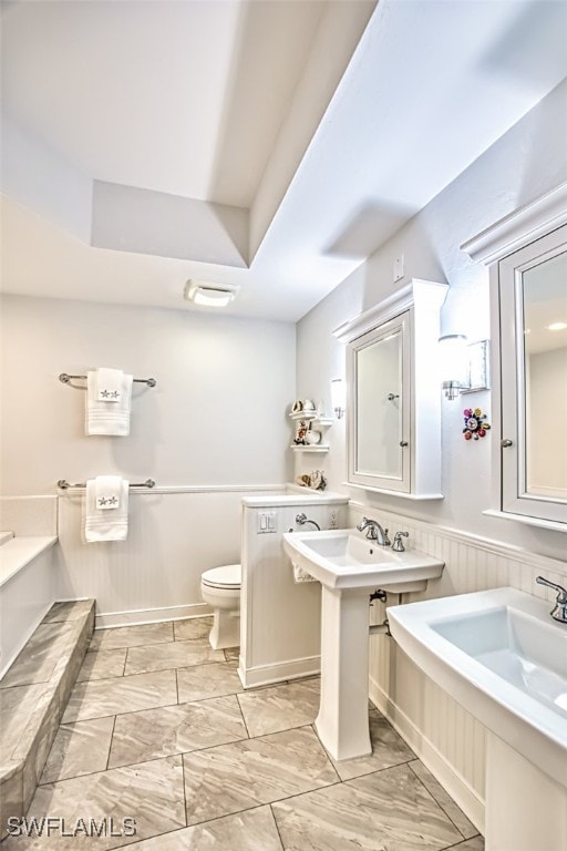 full bath featuring toilet, a bath, two sinks, and wainscoting