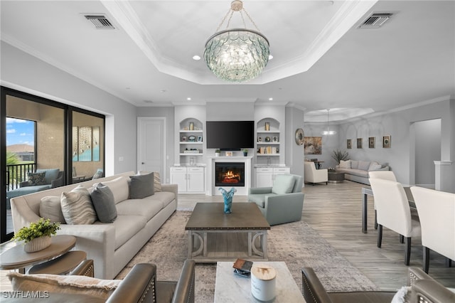 living room featuring built in features, ornamental molding, a chandelier, a tray ceiling, and light wood-type flooring