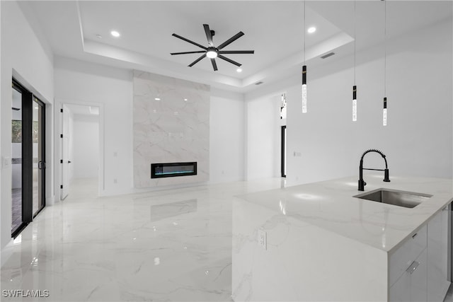 kitchen featuring an island with sink, sink, pendant lighting, a raised ceiling, and light stone counters