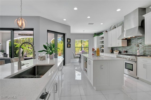 kitchen featuring stainless steel stove, hanging light fixtures, sink, light stone counters, and wall chimney range hood