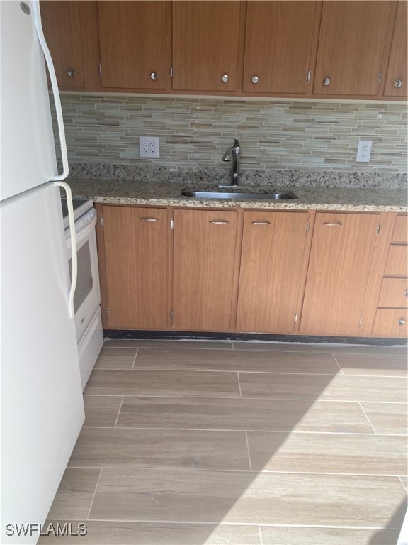 kitchen with brown cabinets, decorative backsplash, a sink, light stone countertops, and white appliances