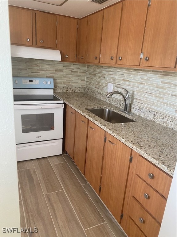 kitchen with electric stove, brown cabinets, backsplash, a sink, and under cabinet range hood