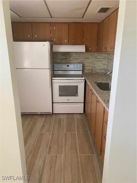 kitchen with under cabinet range hood, white appliances, a sink, decorative backsplash, and brown cabinetry