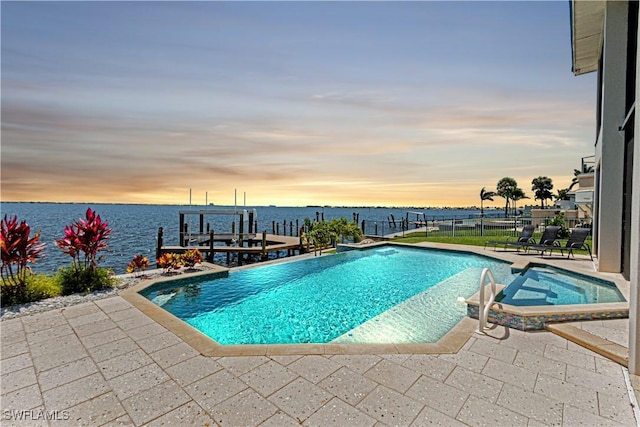 pool at dusk featuring a pool with connected hot tub, a water view, a patio area, fence, and a dock