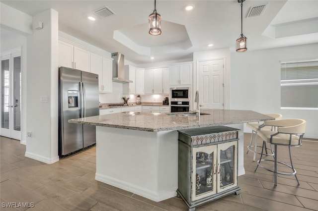 kitchen with wall chimney exhaust hood, stainless steel appliances, white cabinets, and a center island with sink