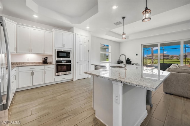 kitchen featuring stainless steel appliances, a tray ceiling, decorative light fixtures, a kitchen island with sink, and sink