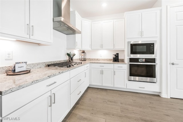 kitchen with wall chimney range hood, white cabinetry, appliances with stainless steel finishes, light stone countertops, and light hardwood / wood-style floors