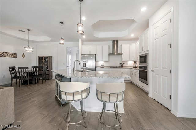 kitchen featuring a tray ceiling, wall chimney exhaust hood, stainless steel appliances, light hardwood / wood-style flooring, and a center island with sink