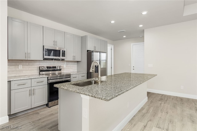 kitchen with backsplash, light wood-type flooring, light stone counters, stainless steel appliances, and an island with sink