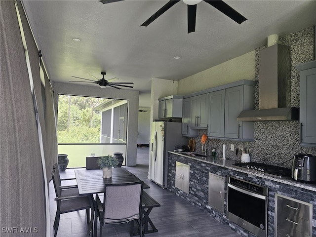 kitchen with black gas cooktop, gray cabinetry, wall chimney range hood, stainless steel oven, and ceiling fan