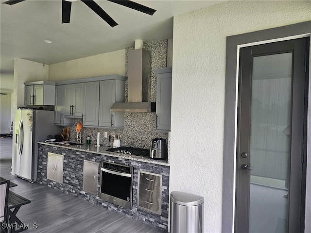 kitchen featuring stone countertops, appliances with stainless steel finishes, decorative backsplash, ceiling fan, and wall chimney range hood