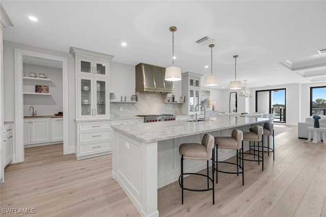 kitchen with light wood-type flooring, wall chimney range hood, white cabinets, and a large island with sink