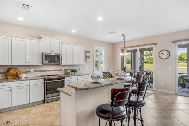 kitchen with a center island with sink, tasteful backsplash, white cabinetry, appliances with stainless steel finishes, and decorative light fixtures