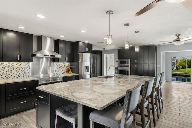 kitchen featuring stainless steel appliances, a sink, a ceiling fan, wall chimney range hood, and backsplash