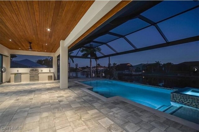 pool at night with glass enclosure, ceiling fan, an outdoor kitchen, and a patio