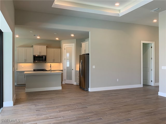 kitchen with light hardwood / wood-style flooring, stainless steel appliances, a raised ceiling, and a kitchen island with sink