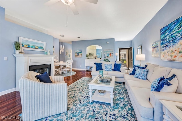 living area featuring arched walkways, a ceiling fan, a glass covered fireplace, wood finished floors, and baseboards