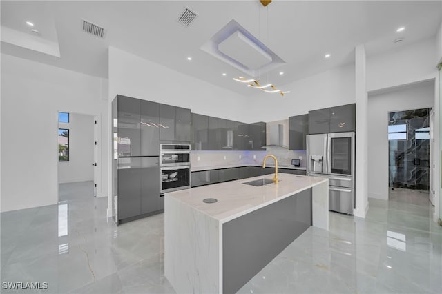 kitchen with gray cabinets, decorative light fixtures, an island with sink, sink, and wall chimney range hood