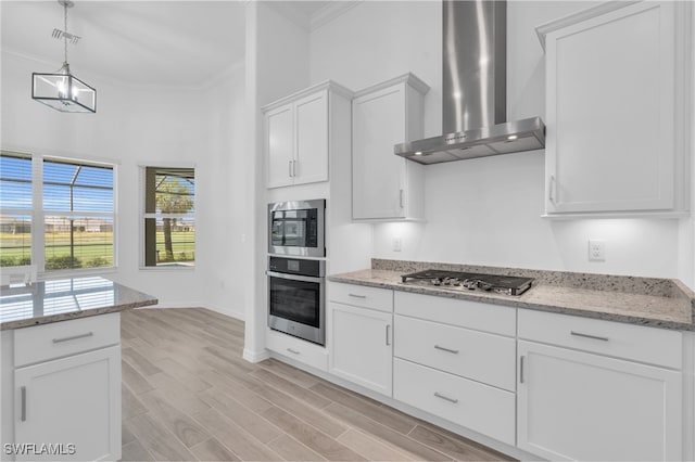 kitchen with crown molding, wall chimney range hood, appliances with stainless steel finishes, white cabinets, and light hardwood / wood-style floors