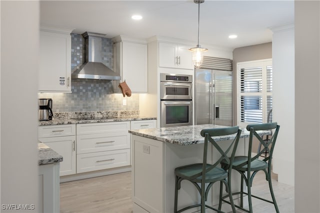 kitchen with appliances with stainless steel finishes, hanging light fixtures, white cabinets, wall chimney exhaust hood, and light wood-type flooring