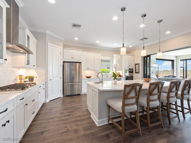 kitchen with appliances with stainless steel finishes, hanging light fixtures, an island with sink, dark hardwood / wood-style flooring, and wall chimney range hood