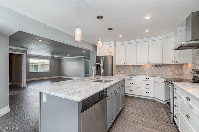 kitchen with an island with sink, ceiling fan, sink, appliances with stainless steel finishes, and wall chimney range hood