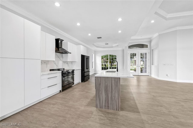 kitchen featuring black appliances, white cabinetry, a kitchen island with sink, a tray ceiling, and wall chimney range hood