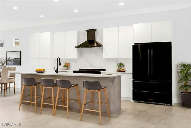 kitchen featuring wall chimney range hood, a kitchen island with sink, white cabinets, black fridge, and decorative backsplash