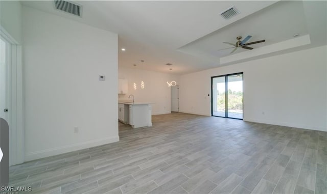 unfurnished living room with light wood-type flooring, ceiling fan with notable chandelier, a tray ceiling, and sink