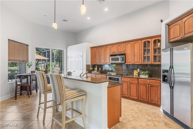 kitchen with dark stone countertops, stainless steel appliances, tasteful backsplash, an island with sink, and decorative light fixtures