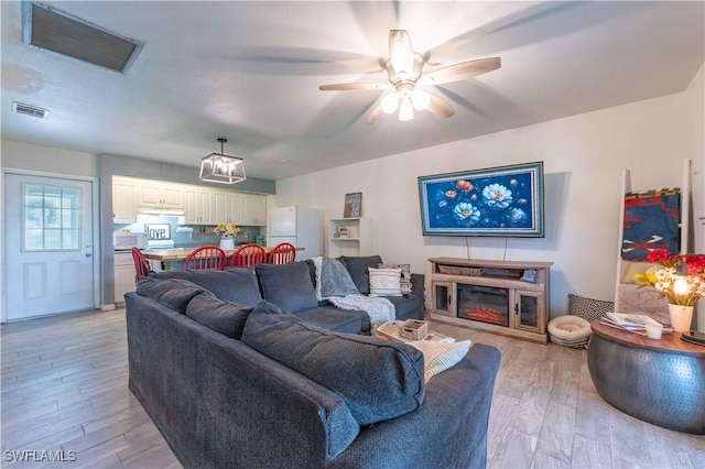 living room with light wood-style floors, visible vents, a ceiling fan, and a glass covered fireplace