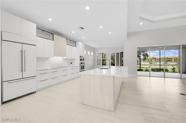 kitchen featuring paneled built in refrigerator, white cabinets, a wealth of natural light, sink, and a kitchen island with sink