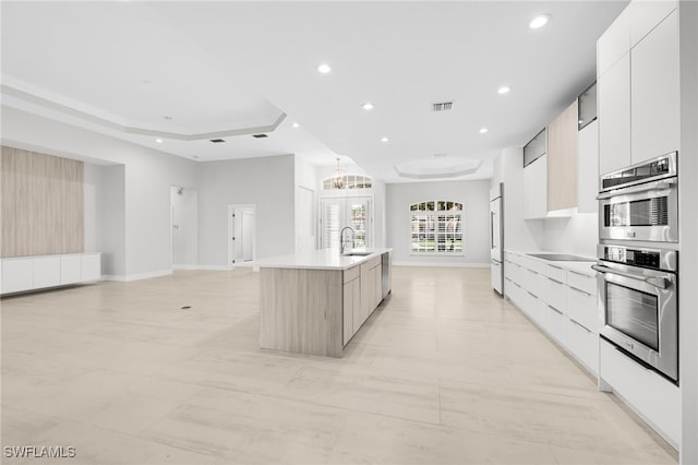 kitchen with double oven, a tray ceiling, sink, white cabinetry, and a kitchen island with sink